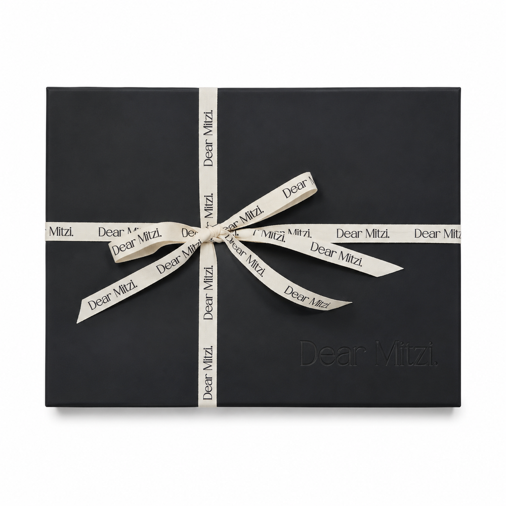 A black rectangular gift box wrapped with a cream-colored ribbon printed with the brand name 'Dear Mitzi.' in black, tied in a bow at the center against a plain white background.