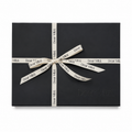 A black rectangular gift box wrapped with a cream-colored ribbon printed with the brand name 'Dear Mitzi.' in black, tied in a bow at the center against a plain white background.