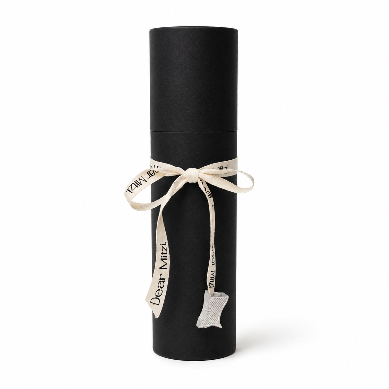 A black cylindrical gift box tied with a cream ribbon printed with the words 'Dear Mirzi' and featuring a small white lace tag, shown on a plain white background.